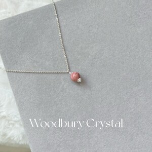 May include: A silver chain necklace with a pink and white gemstone pendant. The pendant is round and has a small, clear crystal on the side. The necklace is on a gray background with the text "Woodbury Crystal" printed below.