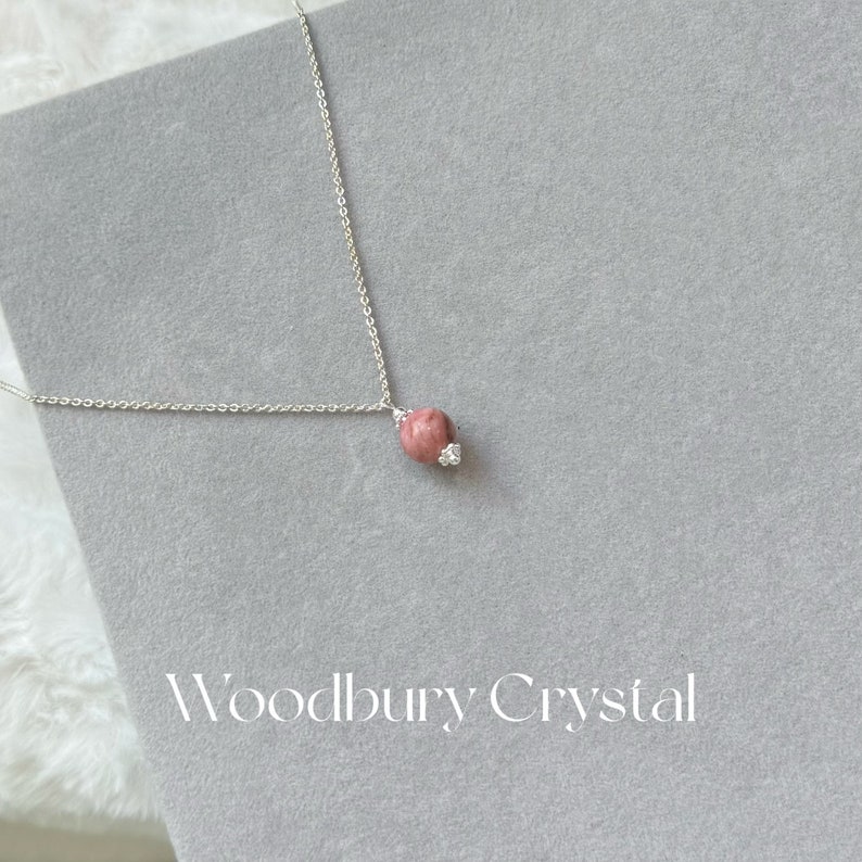 May include: A silver chain necklace with a pink and white gemstone pendant. The pendant is round and has a small, clear crystal on the side. The necklace is on a gray background with the text "Woodbury Crystal" printed below.