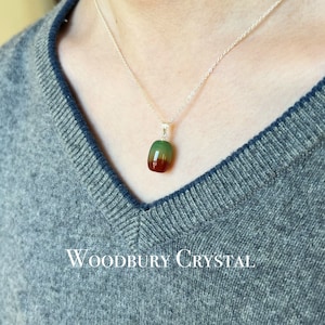 May include: A silver chain necklace with a green, brown, and red gemstone pendant. The pendant is a smooth, rounded shape. The necklace is on a person's chest, over a gray sweater. The text "WOODBURY CRYSTAL" is visible at the bottom of the image.