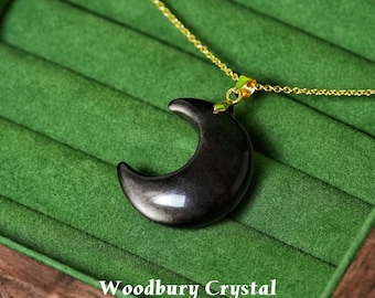 May include: A black crescent moon pendant necklace with a gold chain. The moon pendant is smooth and shiny, with a subtle metallic sheen. The necklace is displayed on a green velvet surface. The text "Woodbury Crystal" is at the bottom.