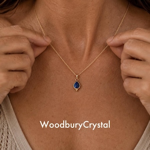 May include: A gold-colored necklace with a teardrop-shaped, dark blue pendant. The pendant has a small gold-colored accent at the bottom. The necklace is displayed on a person's chest. The text "WoodburyCrystal" is at the bottom of the image.