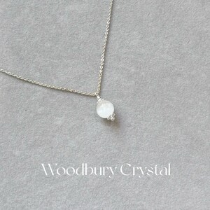 May include: A silver chain necklace with a white moonstone pendant. The pendant is round and has a delicate design. The necklace is on a gray background.