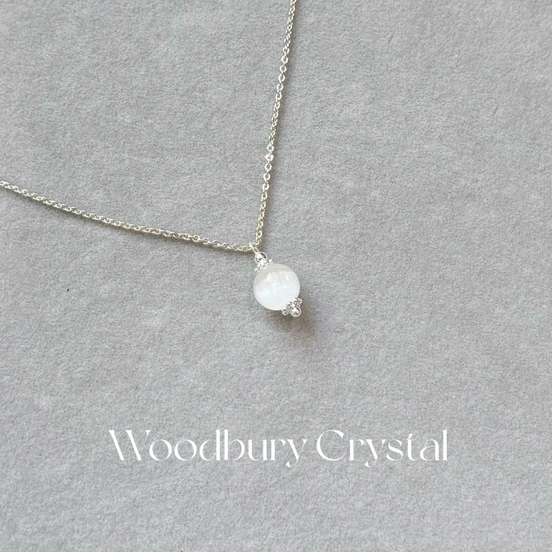 May include: A silver chain necklace with a white moonstone pendant. The pendant is round and has a delicate design. The necklace is on a gray background.
