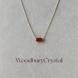 May include: A silver chain necklace with a single, small, rectangular, red carnelian gemstone pendant. The pendant is centered on the chain. The text "Woodbury Crystal" is printed in purple below the necklace.