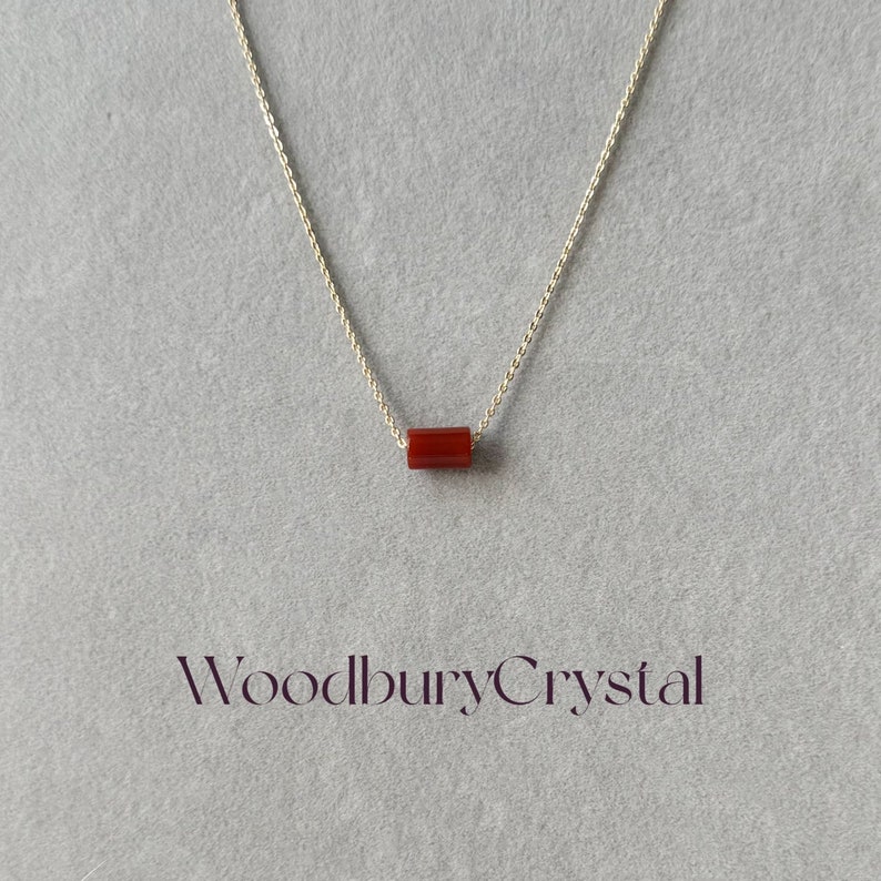 May include: A silver chain necklace with a single, small, rectangular, red carnelian gemstone pendant. The pendant is centered on the chain. The text "Woodbury Crystal" is printed in purple below the necklace.