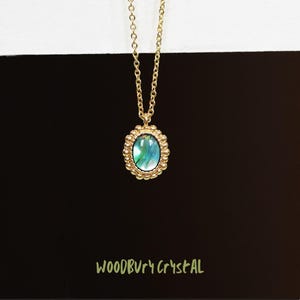 May include: A gold chain necklace with a green and blue oval pendant set in a gold beaded frame. The pendant has a pearlescent finish. The text "WOODBURY CRYSTAL" is visible at the bottom of the image.