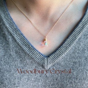 Puede incluir: Un collar de cadena de oro con un pequeño colgante de cristal iridiscente. El cristal está envuelto en alambre de oro y tiene un tono rosa claro y azul.