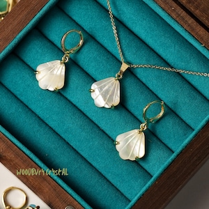 Pode incluir: Um conjunto de brincos de ouro em forma de concha e um colar com um pingente de concha combinando. As joias são exibidas em uma caixa de madeira com um forro de veludo turquesa. O texto "WOODBURY CRYSTAL" é visível na caixa.