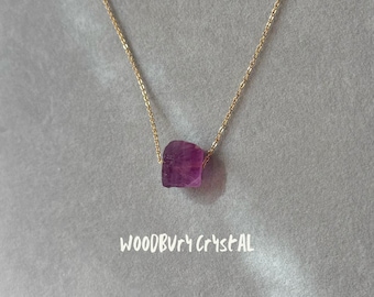 May include: A delicate gold chain necklace with a single, faceted, purple crystal pendant. The pendant is a natural, irregular shape and hangs from the chain.