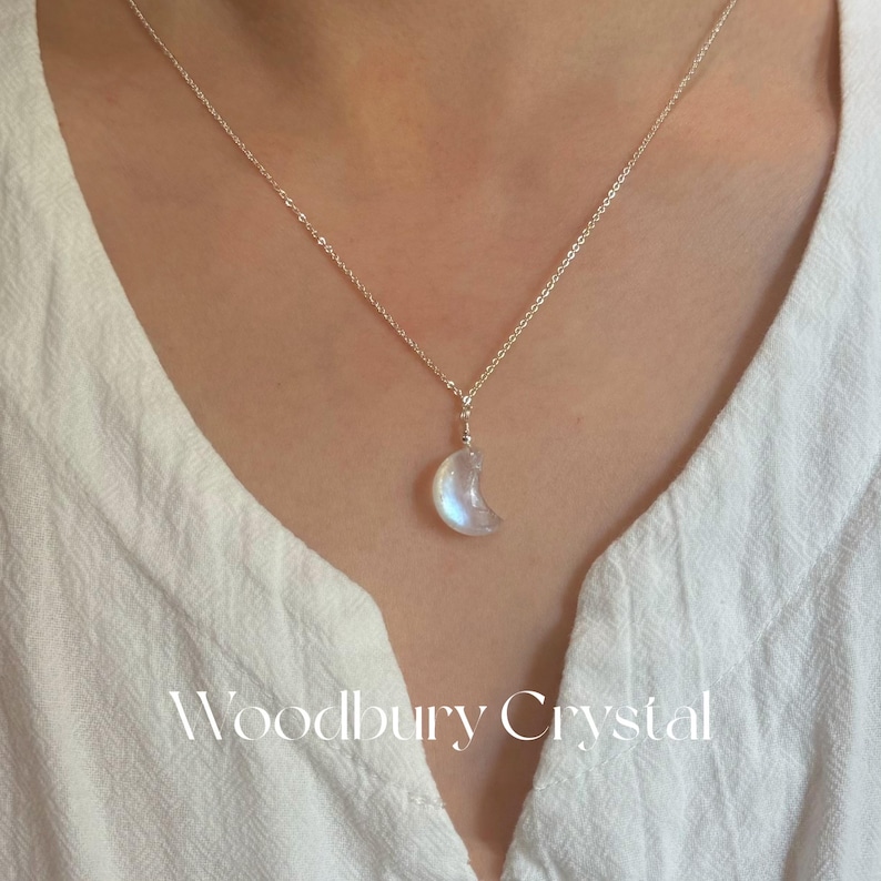 May include: A silver chain necklace with a crescent moon-shaped rainbow moonstone pendant. The pendant is a light blue color with a white background. The necklace is on a model wearing a white shirt. The text "Woodbury Crystal" is visible at the bottom of the image.