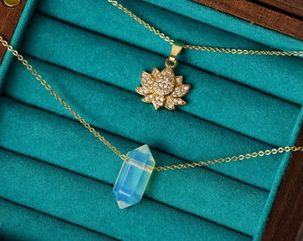 May include: Two gold necklaces in a teal velvet jewelry box. One necklace features a crystal pendant in the shape of a lotus flower. The other necklace features a crystal pendant in the shape of a long, thin, rectangular prism. The text "WOODBURY CRYSTAL" is visible at the bottom of the image.