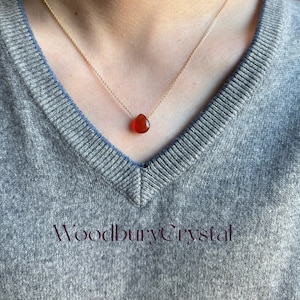 May include: A gold chain necklace with a teardrop-shaped carnelian gemstone pendant. The necklace is worn by a person with a grey sweater.