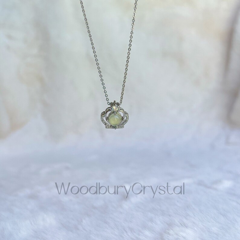 Puede incluir: Un collar de plata con un colgante de corona que presenta una piedra preciosa verde claro. La corona est&aacute; adornada con peque&ntilde;as piedras transparentes.