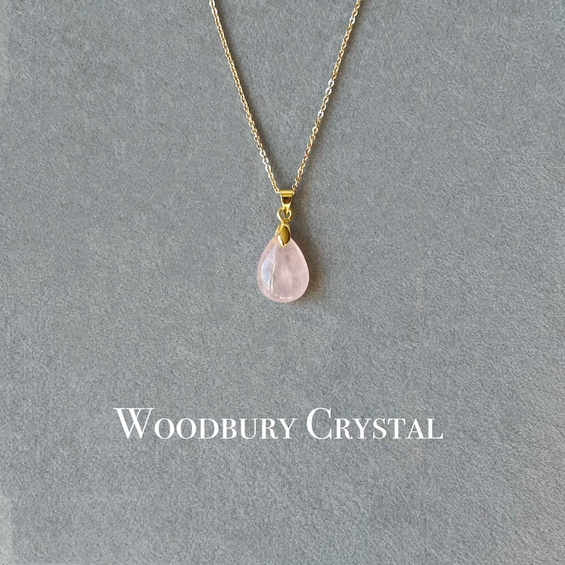 May include: A delicate gold chain necklace with a light pink teardrop-shaped pendant. The pendant is made of rose quartz and has a subtle sparkle. The necklace is labeled "Woodbury Crystal".