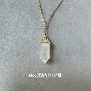 May include: A gold chain necklace with a clear quartz crystal pendant. The pendant is shaped like a point and has a gold wire wrapped around the top. The chain has small gold beads.