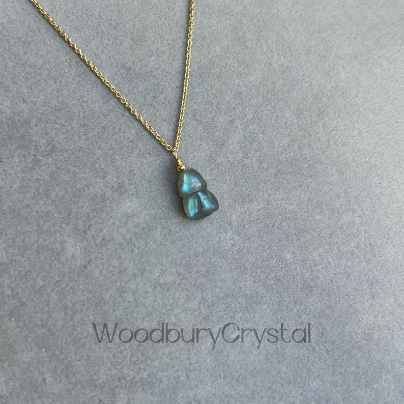 May include: A gold chain necklace with a blue-green labradorite pendant in the shape of a teardrop. The pendant has a shimmery, iridescent quality. The necklace is on a gray background with the text "WoodburyCrystal" printed below.