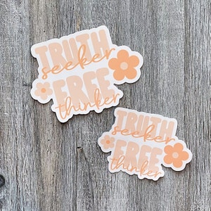 Op de afbeelding: Twee stickers met de woorden "TRUTH seeker FREE thinker" in een retro lettertype, met perzik kleurige bloemen. De stickers hebben een witte achtergrond en een perzik- en lichtoranje kleurenschema. De stickers liggen op een houten oppervlak.