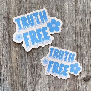Op de afbeelding: Twee stickers met de woorden "TRUTH seeker FREE thinker" in licht- en donkerblauw, met bloemaccenten. De stickers liggen op een houten oppervlak.