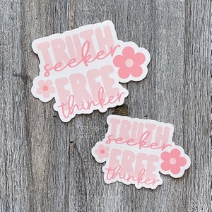 Op de afbeelding: Twee lichtroze stickers met de woorden "TRUTH seeker FREE thinker" in een retro lettertype. Elke sticker heeft een bloemafbeelding. De stickers liggen op een verweerd houten oppervlak.