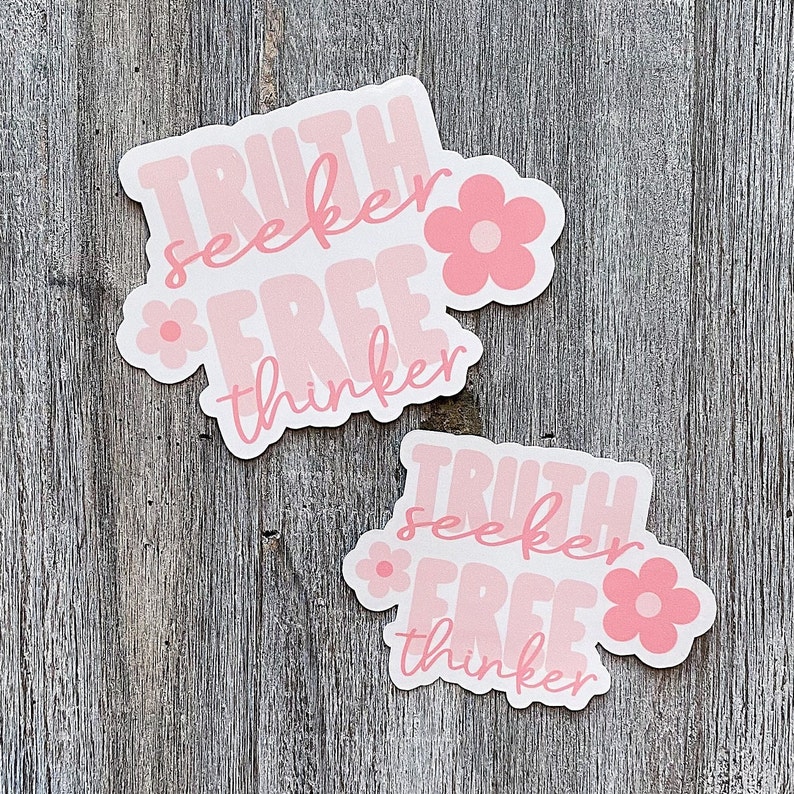 Op de afbeelding: Twee lichtroze stickers met de woorden "TRUTH seeker FREE thinker" in een retro lettertype. Elke sticker heeft een bloemafbeelding. De stickers liggen op een verweerd houten oppervlak.