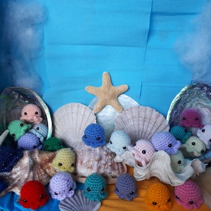 May include: A collection of crocheted octopus plushies in various colours, including red, blue, green, purple, and pink. The plushies are arranged in a seashell display with a starfish on top.