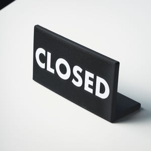 May include: A black sign with the word "CLOSED" in bold white letters. The sign is rectangular and stands upright on a small base. The background is white.