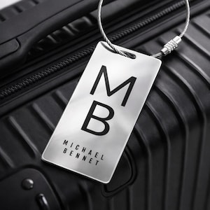May include: A silver luggage tag with the initials "MB" in bold black letters, attached to a black suitcase. The tag also displays the name "MICHAEL BENNET" in a smaller font. The suitcase has a textured surface.