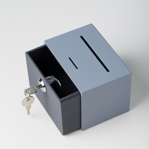 May include: A gray and dark gray lockbox with a key in the lock. The box has a slot on the top and a drawer that is partially open. The key is silver.