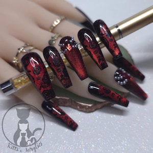May include: Long, coffin-shaped acrylic nails in a deep red hue with black floral designs. Some nails are adorned with small, silver-colored beads. The nails are from Kitti's Acrylics.