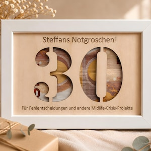 Könnte beinhalten: Ein weißer rechteckiger Holzrahmen mit einem hellen Holzeinsatz, in den die Zahl 30 ausgeschnitten ist. Der Text Steffans Notgroschen! und Für Fehlentscheidungen und andere Midlife-Crisis-Projekte ist in das Holz eingraviert, wobei Euro-Banknoten hinter dem Ausschnitt sichtbar sind.