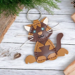 May include: A brown and tan leather cat keychain with blue eyes and white whiskers. The cat is sitting on a white wooden surface.