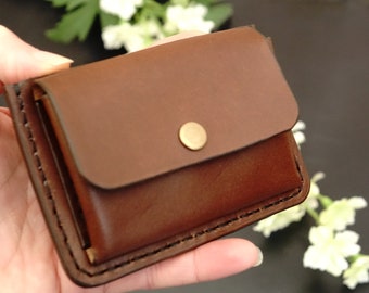 Puede incluir: Una cartera de cuero marrón con cierre de solapa y un botón de latón. La cartera tiene un borde cosido y está doblada por la mitad.
