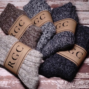 Könnte beinhalten: Ein Stapel von sechs Paar Wollsocken in verschiedenen Brauntönen, Grau- und Schwarztönen. Jedes Paar ist in braunes Papier eingewickelt, auf dem der Text "RGC" gedruckt ist.