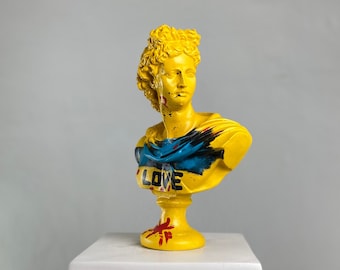 May include: A yellow painted bust sculpture of a woman with a blue and black painted design on the chest. The bust is on a white pedestal with a decorative scroll design. The bust has the word "LOVE" painted on the chest.