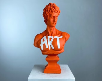 May include: Orange painted bust sculpture of a man with curly hair on a white pedestal. The bust has the word "ART" written across the chest in white paint.