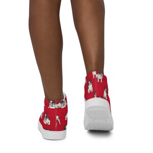 Puede incluir: Un par de zapatillas blancas de lona de caña alta con fondo rojo y un patrón de bulldogs franceses. Las zapatillas las lleva puestas una persona de piel morena.