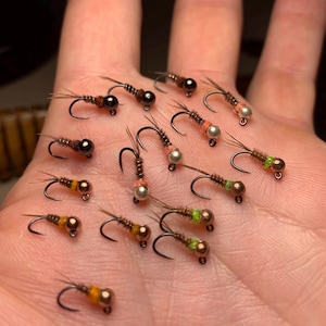 May include: A collection of 17 fly fishing lures with different colored beads and feathers. The lures are arranged on a hand.
