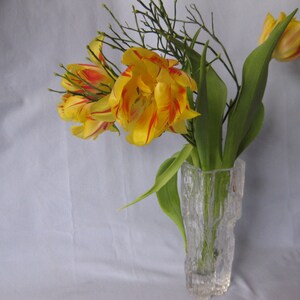 Könnte beinhalten: Eine klare Glasvase mit einem strukturierten, rindenartigen Design enthält einen Strauß gelber und roter Tulpen. Grüne Blätter und dünne, zweigartige Stiele ergänzen das Arrangement. Die Blumen blühen in voller Pracht mit leuchtenden Farben.