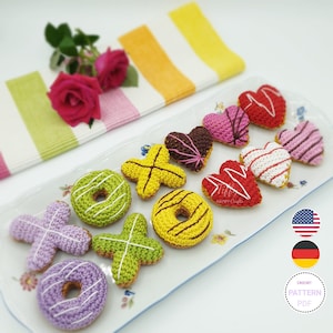 Könnte beinhalten: Eine Sammlung gehäkelter Donuts und Herzen auf einem weißen Teller mit Blumenmustern. Die Donuts sind in verschiedenen Farben, darunter grün, gelb und lila. Die Herzen sind rot, rosa und braun. Der Teller liegt auf einer gestreiften Tischdecke.