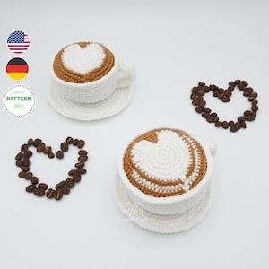 May include: Two crocheted coffee cups with a heart-shaped design in the foam. The cups are white with brown foam and are sitting on white saucers. There are coffee beans arranged in a heart shape around each cup.