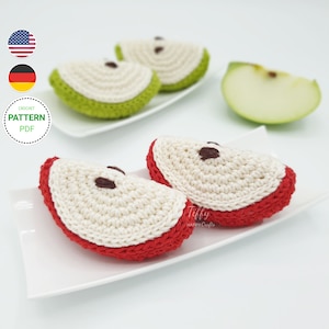 Puede incluir: Dos rodajas de manzana de ganchillo en un plato blanco. Una rodaja es roja y la otra es verde. Las rodajas de manzana están hechas con hilo blanco y tienen centros marrones. El plato está sobre una superficie blanca.