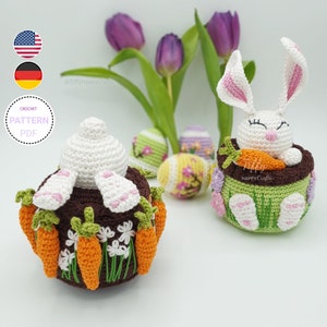 May include: Crochet pattern PDF for making two Easter bunny amigurumi toys. One bunny is sitting in a pot with carrots and the other is standing with carrots around its base. Both bunnies are white with pink ears and noses. The pattern is available in English and German.