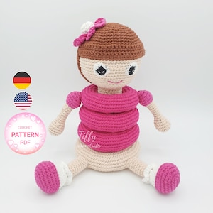 Può includere: Modello a uncinetto PDF per un giocattolo di bambola impilabile rosa e marrone. La bambola ha un fiore sulla testa e indossa un vestito rosa con tre anelli intorno alla vita. La bambola è seduta su una superficie bianca.