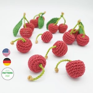 Puede incluir: Un patrón de crochet para hacer cerezas rojas en miniatura con tallos y hojas verdes. El patrón está disponible para descargar en PDF. La imagen muestra varias cerezas de crochet sobre un fondo blanco.