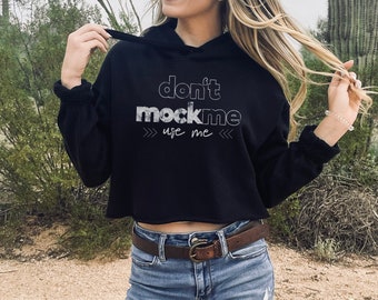May include: A black cropped hoodie with the text "don't mock me use me" in white lettering. The hoodie is worn by a person with long blonde hair and sunglasses.