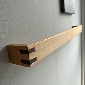 Może przedstawiać: Drewniana półka z ciemnym drewnianym wkładką na rogach. Półka jest zamontowana na jasnoszarej ścianie.