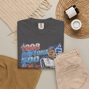 Peut inclure: Un t-shirt &agrave; col rond gris anthracite avec une impression graphique du Daytona 500 de 1998, repr&eacute;sentant un pilote de course portant des lunettes de soleil et une combinaison, avec des &eacute;clairs bleus et un motif de drapeau &agrave; damier.