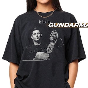 May include: A black short-sleeve t-shirt featuring a black and white graphic of a man in a jacket, a large boot print, and the text "bitch." printed in a small, simple font above the graphic. The shirt has a crew neckline and a loose fit.