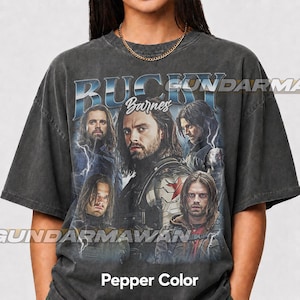 May include: A charcoal gray short-sleeve t-shirt featuring a graphic print of Bucky Barnes with the text Bucky Barnes. The design includes multiple portraits of the character, lightning effects, and a metallic-style font. The shirt has a crew neck.