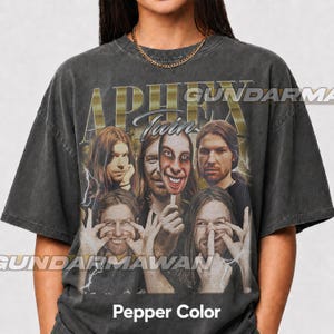May include: A charcoal gray short-sleeve t-shirt featuring a graphic print of Aphex Twin. The design includes multiple portraits of the artist, including one holding a mask, with the text "APHEX" and "Twin" in a gold-toned font and lightning accents.